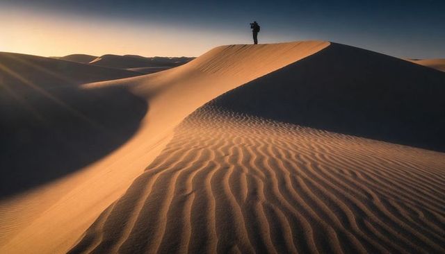 Solitary photographer standing on sand dune at sunset capturing dramatic desert silhouette