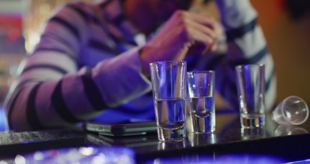 A person sitting in a dimly lit bar or nightclub with three empty shot glasses in front of them. This can be used in content related to nightlife, relaxation, socializing, or articles about responsible drinking.
