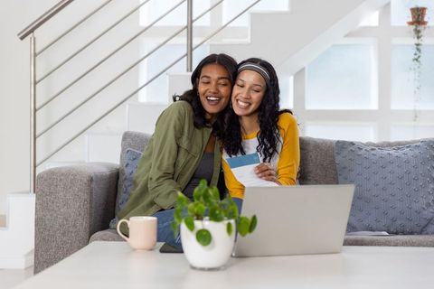 Smiling diverse mother and teenage daughter choosing paint swatches on sofa with laptop