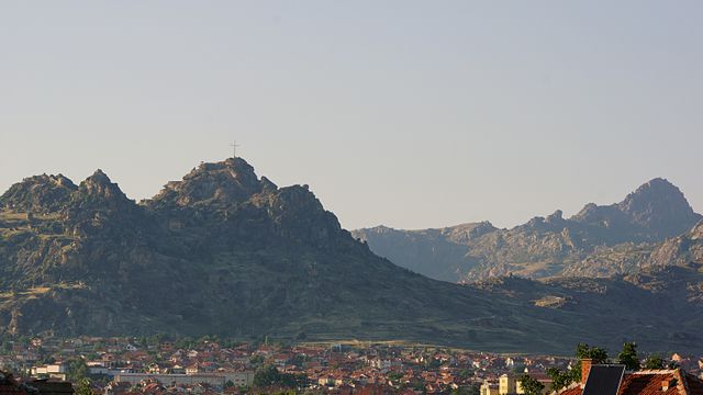 This serene landscape depicts a village nestled at the base of rugged mountains, with a large cross perched on a prominent rocky peak, illuminated by the soft evening light. Ideal for travel guides, tourism advertisements, and nature-inspired projects highlighting peaceful rural life or religious landmarks.