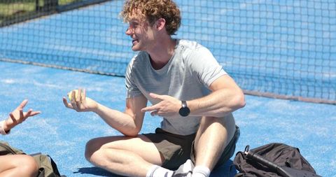 Smiling man sitting cross-legged on blue tennis court holding yellow ball with partner