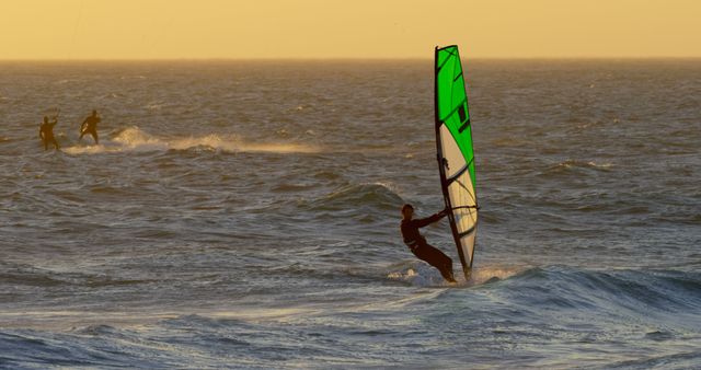 Windsurfer riding waves in the ocean during a vibrant sunset. The sunlight illuminates the scene with a pleasant glow, enhancing the sense of adventure and relaxation. Perfect for marketing materials related to water sports, beach vacations, outdoor activities, and adventure sports promotions.