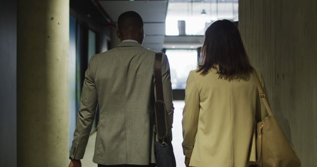 Business professionals seen from the back walking down a modern office corridor. Both are in professional attire and carrying bags, suggesting a productive workday. This can be used to convey themes of teamwork, business environment, collaboration, and a modern office setting.