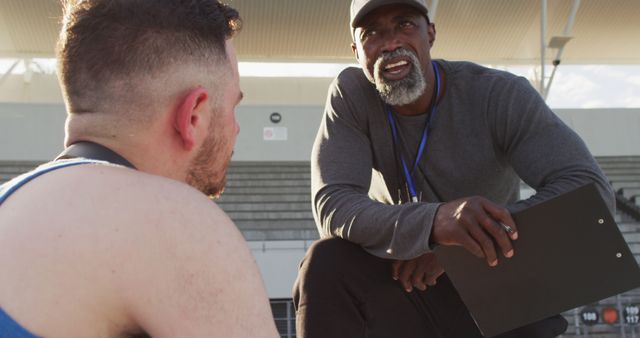 African american male coach and caucasian male athlete sitting and talking. professional runner training at sports stadium.