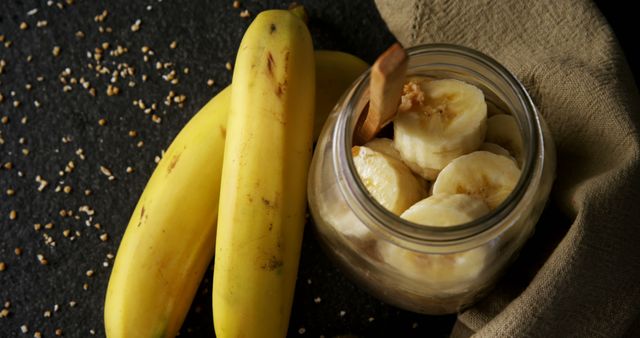 This photo captures fresh sliced bananas in a clear jar accompanied by whole bananas on a dark textured surface. The wooden spoon and seeds scattered around add a rustic touch. Ideal for content related to healthy eating, nutrition, fruit snacks, cooking, and recipes.