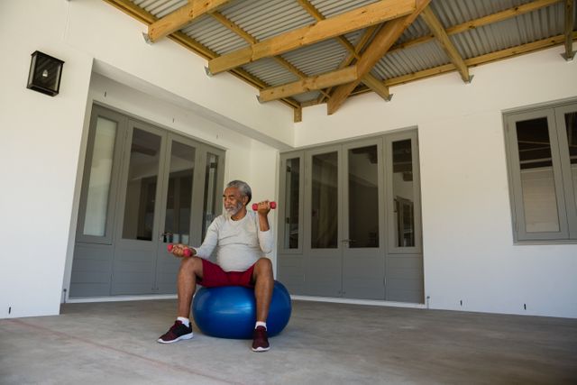 Senior man sitting on exercise ball, lifting dumbbells in porch area. Ideal for promoting senior fitness, active lifestyle, home workouts, and healthy aging. Suitable for use in health and wellness articles, fitness blogs, and retirement living promotions.