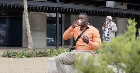 Young african american man sipping coffee and eating sandwich while sitting on urban bench