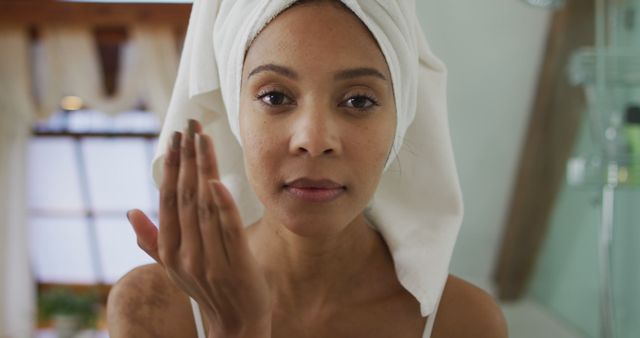 Woman with towel wrapped hair standing in bathroom, applying skincare product. Ideal for use in advertisements or blog posts about skincare routines, personal care tips, beauty tutorials, and health and wellness content.