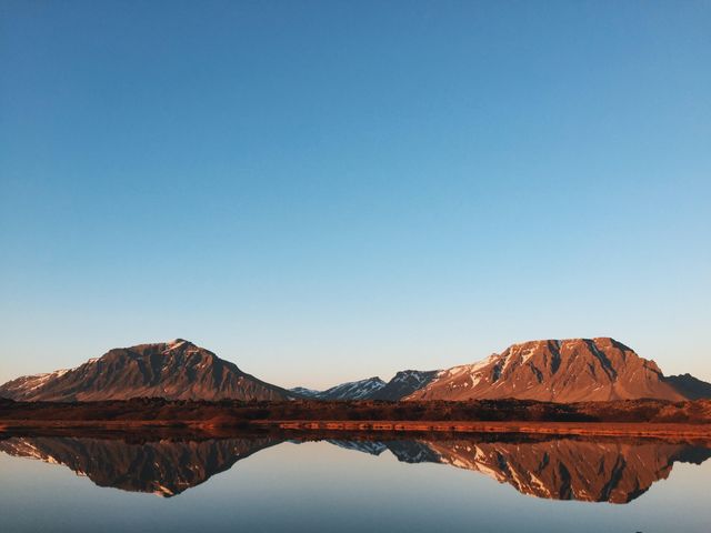 Perfect for use in travel brochures, nature blogs, and meditation app backgrounds, this image captures a stunning mountain landscape reflected perfectly on a calm lake during sunset. The serene and tranquil environment highlighted by the clear blue sky and snowy peaks provides an ideal representation of untouched natural beauty.