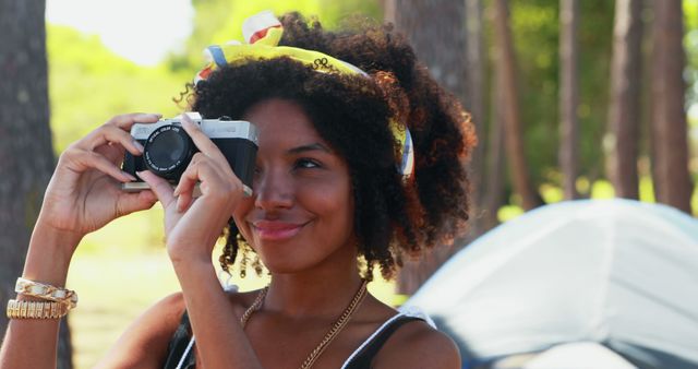 A young African American woman is capturing memories with a vintage camera, with copy space. Her joyful expression and casual camping setting evoke a sense of adventure and leisure.