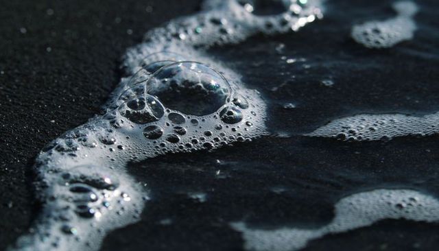 Shimmering sea foam bubbling on black sand shore, iridescent bubbles macro close-up