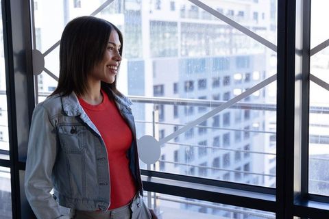 Asian woman smiling on highrise balcony overlooking urban skyline in denim jacket