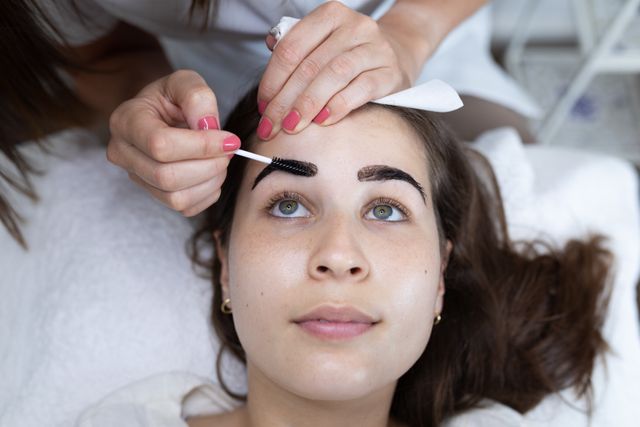 Beautician applying dye to eyebrows of a female client lying down in a beauty salon. Ideal for use in articles or advertisements related to beauty treatments, salon services, cosmetology, and self-care routines.