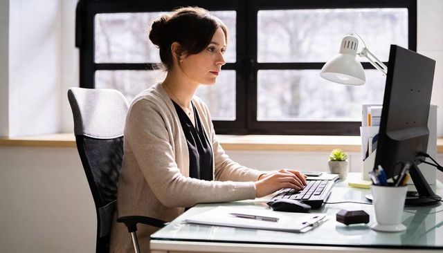 Female professional typing at minimalist glass desk by window, modern office daylight workspace