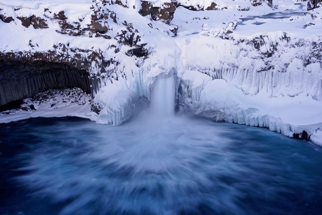 Majestic winter waterfall surrounded by snow and ice, falling into a pool of vibrant blue water. Perfect for travel blogs, nature websites, winter-themed advertising, and artistic wall décor emphasizing natural beauty and tranquility.