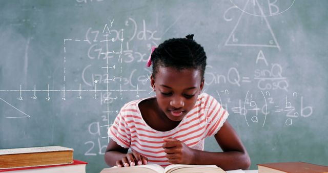Young African American girl focusing on studying math with chalkboard in background covered with equations and graph drawings. Ideal for educational content, school resources, academic websites, textbooks, and classroom materials promoting STEM education.