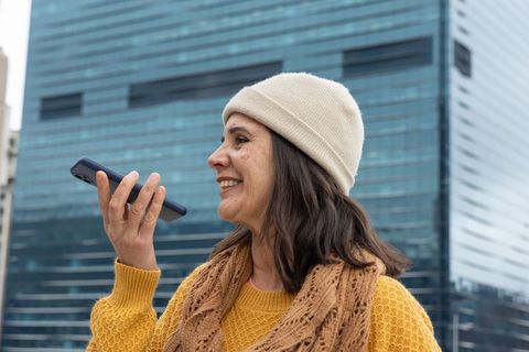 Smiling middle-aged woman using smartphone voice message in urban high-rise backdrop