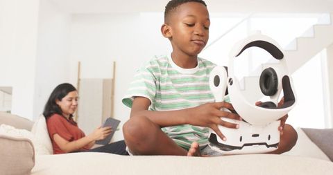 Young boy exploring VR headset on sofa while woman using tablet in bright modern home