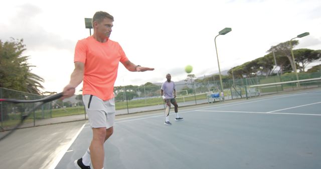 Two men wearing casual sportswear practicing tennis on an outdoor court. One man in orange shirt hitting the ball, with a focus on technique and teamwork. Ideal for content related to sports, fitness, teamwork, exercises, and healthy lifestyle.