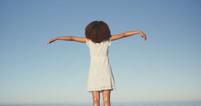 Back view of biracial woman raising hands and standing against blue sky. Summer, relaxation, vacation, happy time.