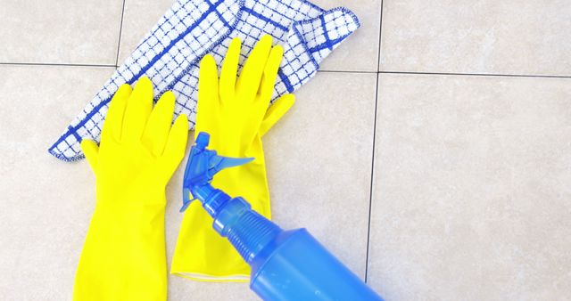 This stock photo shows a pair of yellow cleaning gloves, a blue and white checkered cloth, and a blue spray bottle placed on a tiled floor. Ideal for use in articles or advertisements about house cleaning, sanitation, hygiene routines, or domestic chores. It can also be used in blogs focused on home care tips, cleaning products, or household maintenance strategies.