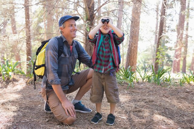 Father and son enjoying a birdwatching adventure in a forest. The boy is looking through binoculars while the father kneels beside him, smiling. Ideal for use in family bonding, outdoor activities, nature exploration, and hiking-related content.