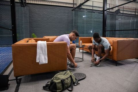 Two male athletes sitting on sofas tying sneakers in sports lounge beside glass court
