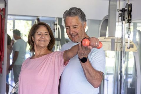 Senior couple exercising with dumbbell in gym, partner supporting shoulder exercise