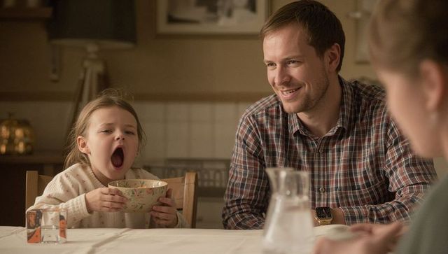Yawning child holding floral bowl while father smiling at cozy morning kitchen table