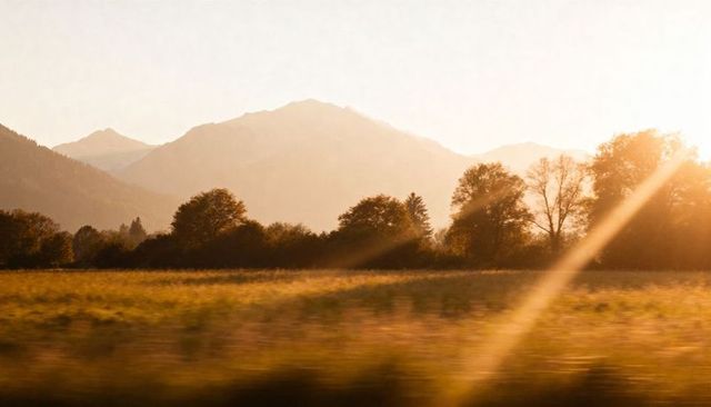 Golden-hour meadow with sun rays, lens flare and hazy mountain ridges at sunset glow