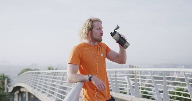 Man seen briefly pausing his morning jog on a bridge to rehydrate with a sports bottle. This can be used for promoting healthy lifestyles, fitness programs, athletic gear, or city's landscape highlighting outdoor exercise spaces.
