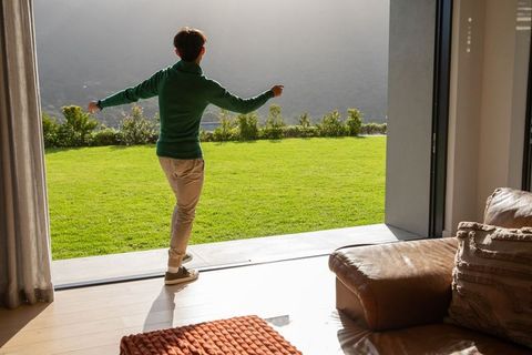 Young man stepping through open sliding door onto sunlit lawn, modern indoor-outdoor living
