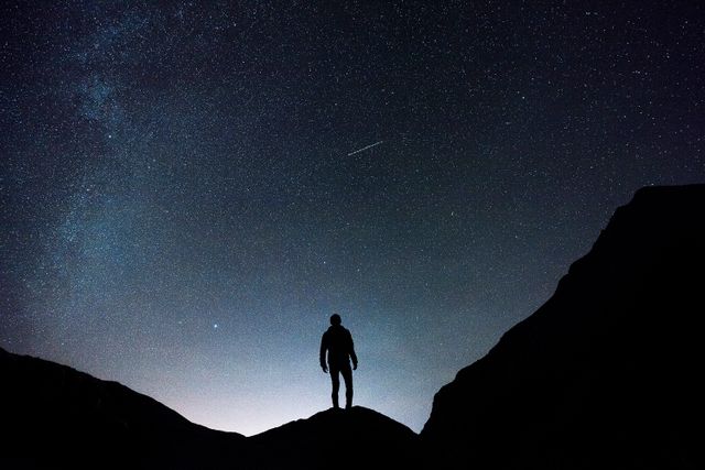 This image shows a person silhouetted against a night sky illuminated by countless stars and the Milky Way, framed by mountain peaks. Ideal for illustrating themes of adventure, solitude, exploration, and the beauty of nature. Perfect for use in travel blogs, outdoor adventure marketing, astronomy articles, and inspirational posters.