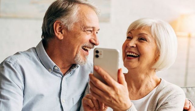 Senior couple laughing and sharing smartphone on couch showing warm connection