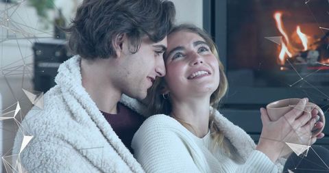 Young couple snuggling by fireplace in sherpa blanket holding mug, cozy winter romance