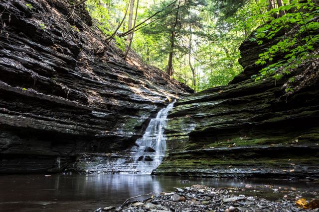 A peaceful waterfall flows through rocky cliffs surrounded by lush green forest. Ideal for use in nature-themed projects, travel brochures, environmental conservation campaigns, or as calming visuals for wellness and mental health content.