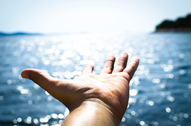 A person's hand is reaching out towards a sunlit ocean with sparkling water in the background. The image portrays themes of travel, adventure, and freedom. It can be used in projects related to summer vacations, natural beauty, personal journeys or motivational messages.