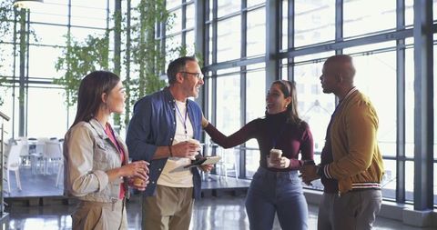 Diverse colleagues chatting and networking over coffee in sunlit atrium with lanyards