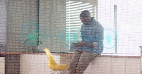 Man leaning on office counter checking smartphone with laptop and yellow chair in sunlit workspace