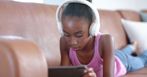 African american teen wearing white headphones using tablet on brown leather sofa