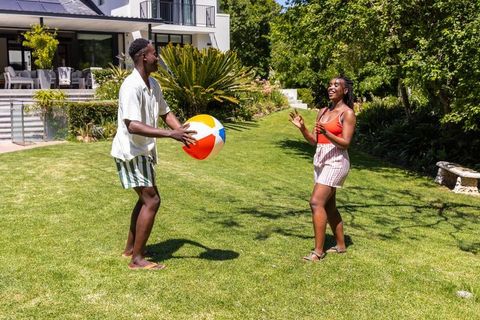 African american couple tossing beach ball on sunny backyard lawn in front of modern house