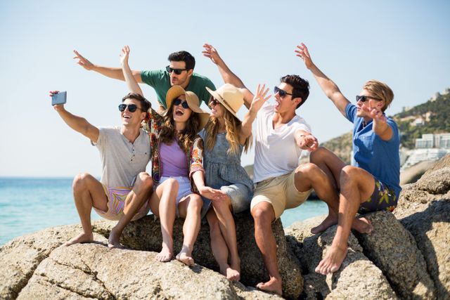 Group of young friends enjoying a sunny day at the beach, taking a selfie while sitting on rock formations. Perfect for use in travel promotions, summer vacation advertisements, social media campaigns, and lifestyle blogs.