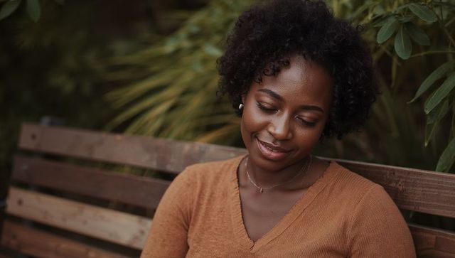 Contemplative woman sitting on wooden bench wearing rust sweater, smiling softly outdoors