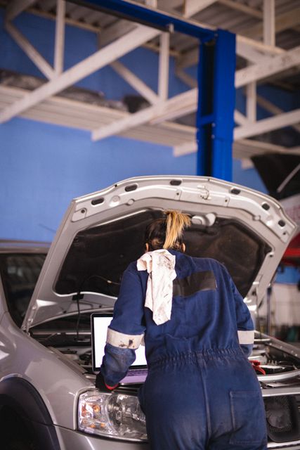 Biracial female car mechanic wearing face mask and overalls, inspecting car, using laptop. independent business owner at car servicing garage during covid 19 pandemic.