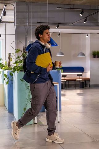 African american teen walking through campus lounge carrying yellow folder, coffee and headphones