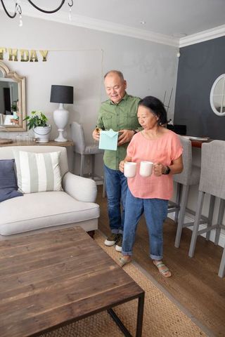 Asian couple celebrating birthday at home holding mugs and opening blue gift bag