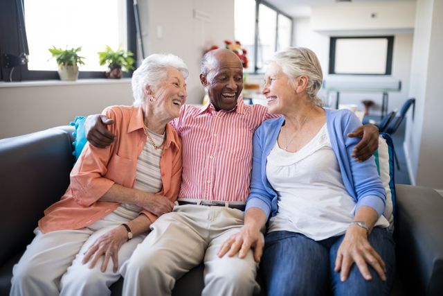 Senior man and two female friends sitting on a sofa, smiling and enjoying each other's company in a nursing home. This image can be used for promoting senior living communities, elderly care services, and social activities for seniors. It highlights themes of friendship, happiness, and the importance of social connections in retirement.