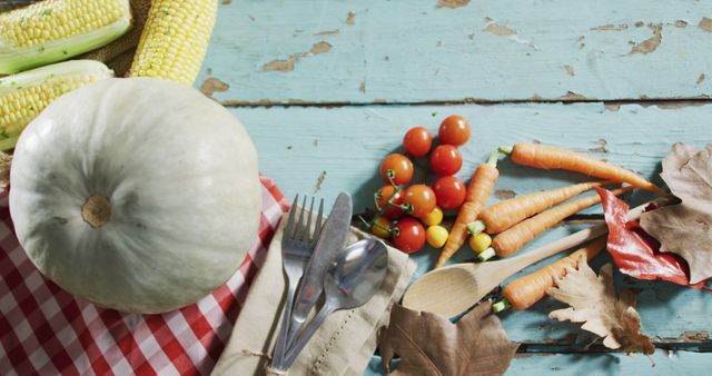 Autumn harvest vegetables, including a white pumpkin, carrots, cherry tomatoes, and corn, are arranged on a rustic blue wooden table. Utensils are placed on a red checkered cloth, and dry leaves enhance the seasonal feel. Perfect for illustrations of organic farming, healthy eating, seasonal recipes, and countryside lifestyle content.