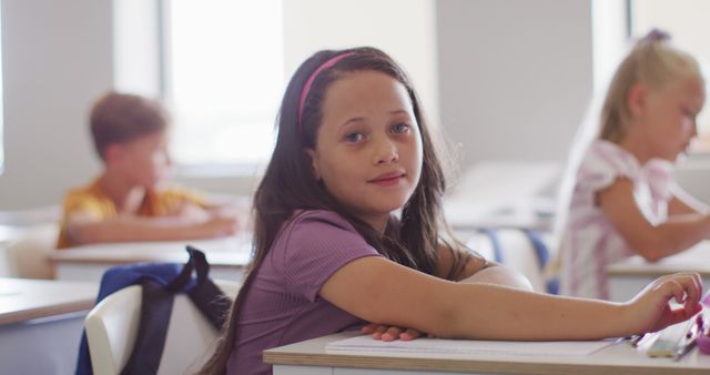 Smiling girl sitting at desk in classroom with classmates in background. Ideal for educational materials, school advertisements, or articles promoting the importance of classroom learning and child development.