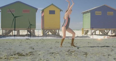 Girl jumping on sunlit beach raising arms beside colorful stilted beach cabins and turbines